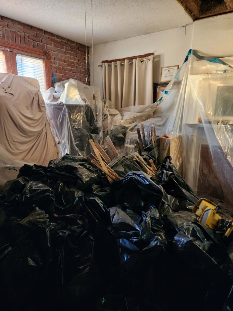A room undergoing renovation is filled with black trash bags and plastic-covered furniture.