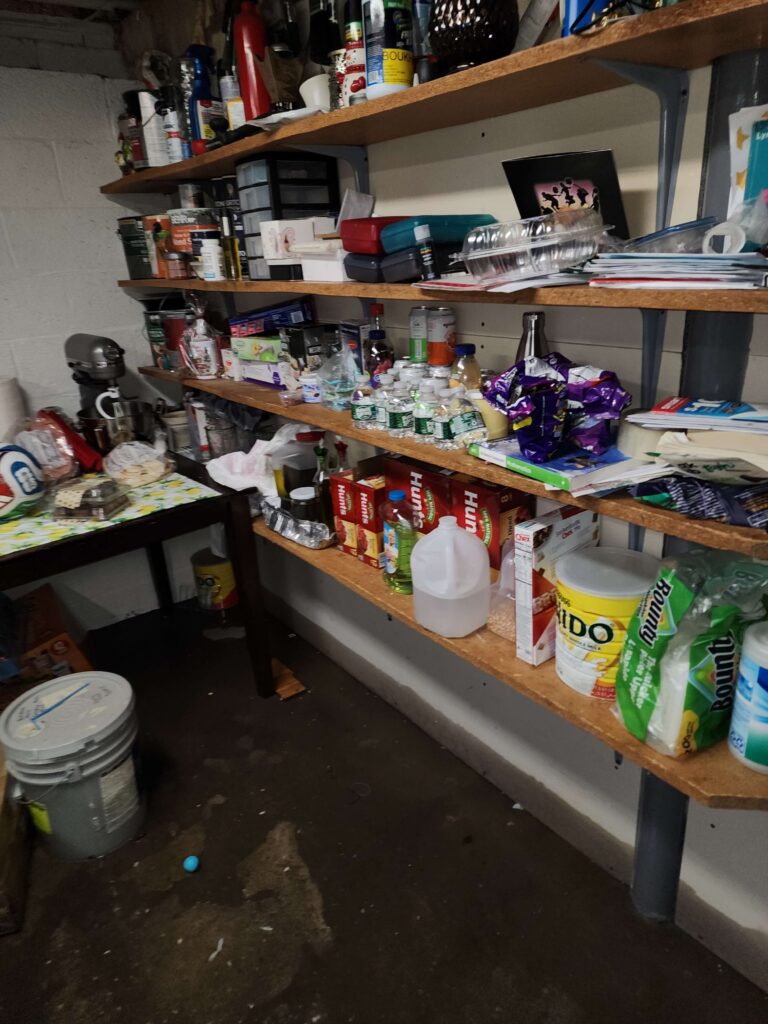 Cluttered wooden shelves in a storage room are packed with food, cleaning supplies, and household