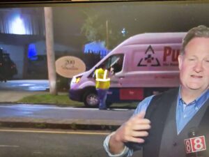 A news reporter gestures while a worker in a safety vest stands by a white service van at