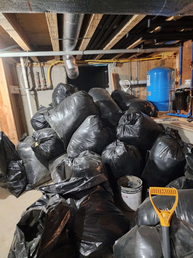 A large pile of black trash bags fills an unfinished basement with exposed pipes and a blue water