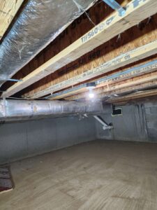 A brightly lit unfinished basement or crawl space featuring exposed wooden joists, ductwork