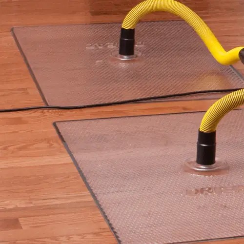 Clear floor drying mats with yellow hoses are suctioned to a wooden floor for water removal.