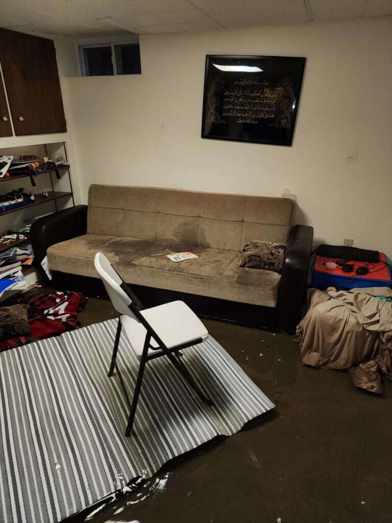 A flooded basement room shows a water-stained sofa, partially submerged rug, and scattered items