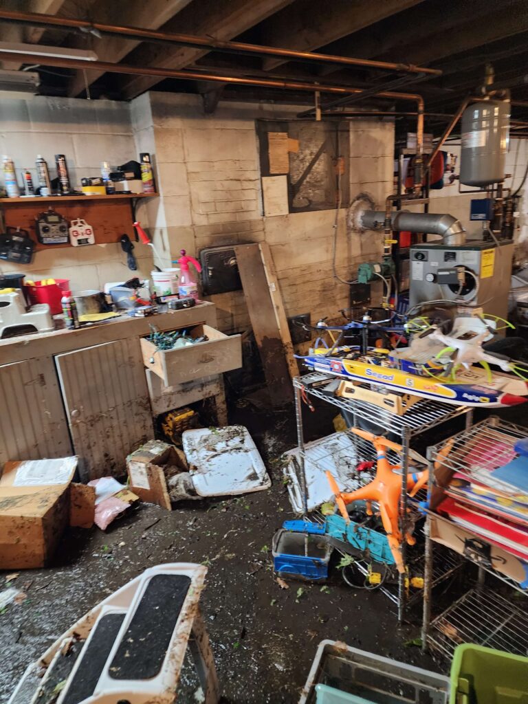 A dark, flooded basement shows water-stained walls, exposed pipes, and scattered debris on