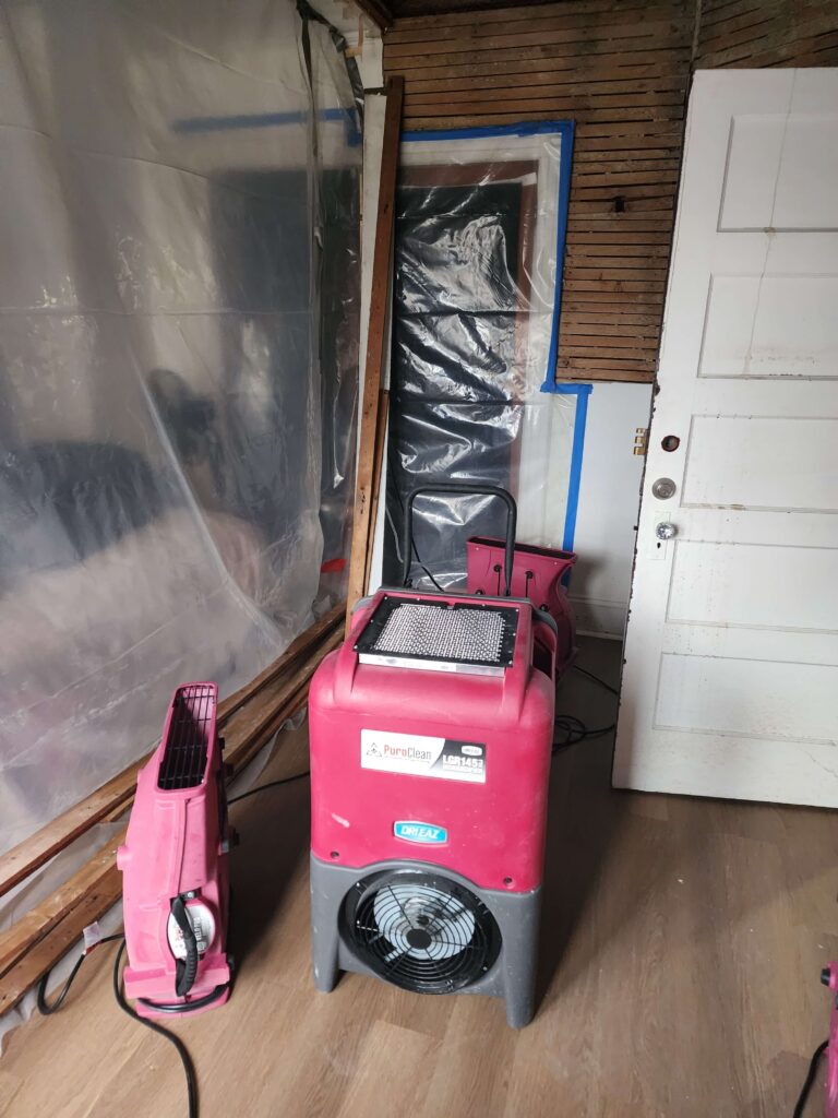 Two red restoration machines sit in a room with exposed lath walls and plastic sheeting. TITLE