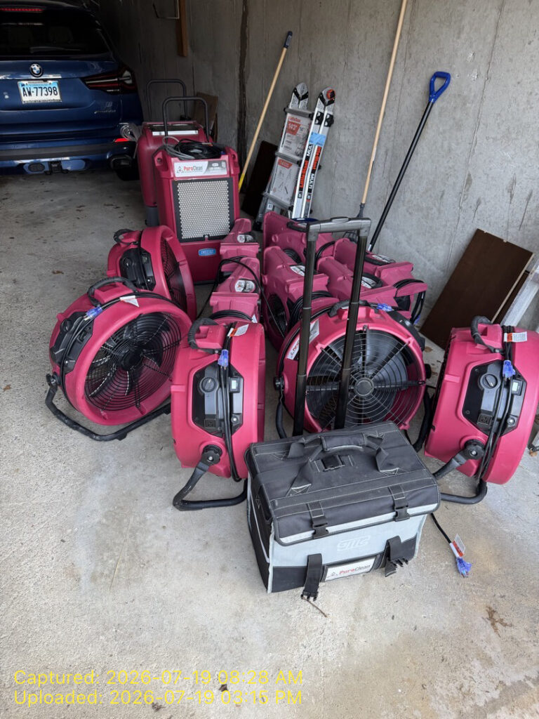 Pink PuroClean water damage restoration equipment, including fans and a dehumidifier, stored in a garage