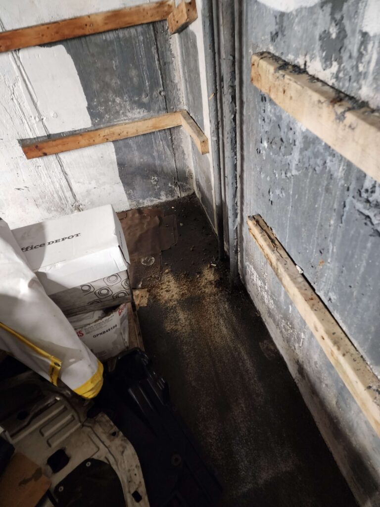 Dirty basement corner with peeling paint, wooden planks, and scattered debris on a dark floor