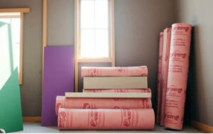 Pink insulation rolls, wood planks, and colorful boards are stacked in a room under construction.