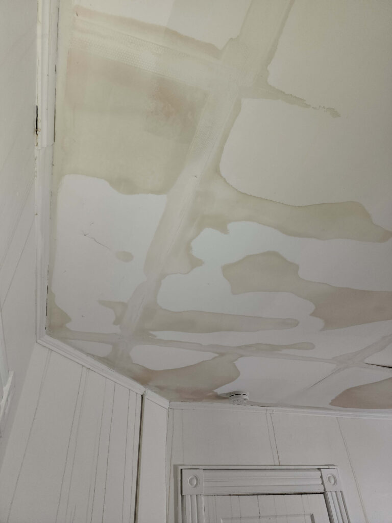 Extensive brown water stains and peeling paint cover a white ceiling and wall, indicating significant water damage.