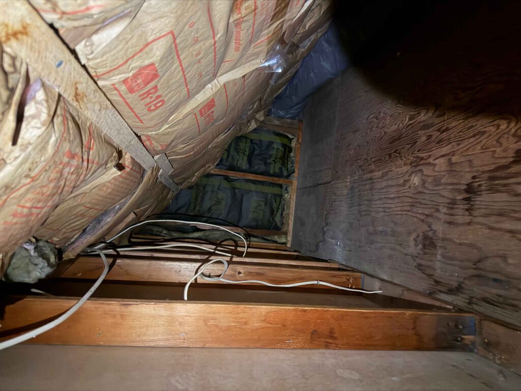 Dark attic interior with R-19 fiberglass insulation, wooden rafters, and exposed electrical