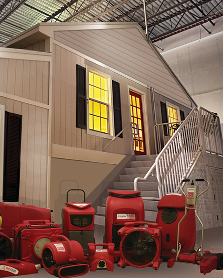 A model house with yellow-lit windows, surrounded by various red water damage restoration equipment.