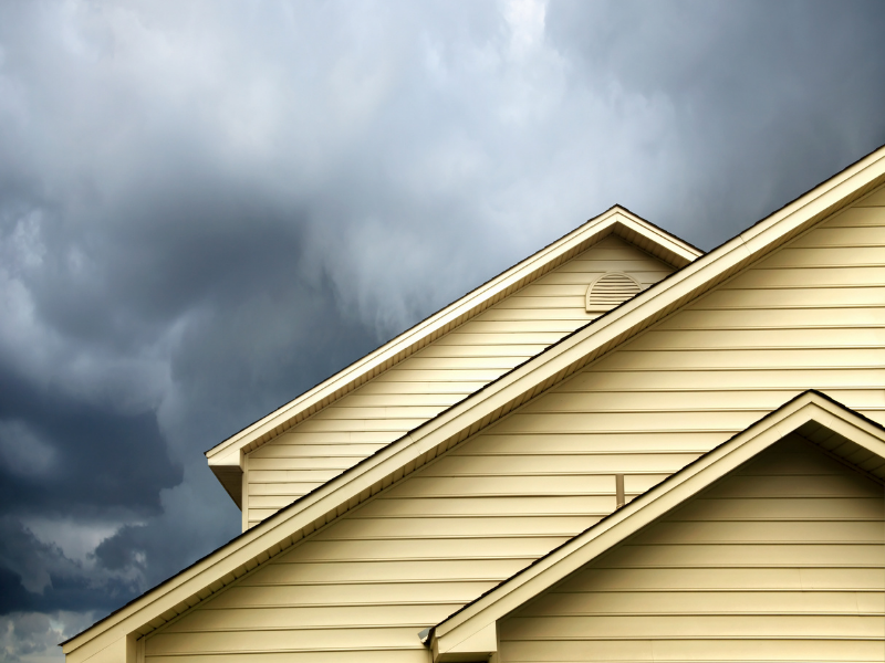 A close-up of a light-colored house under dark storm clouds, highlighting potential water damage risks.