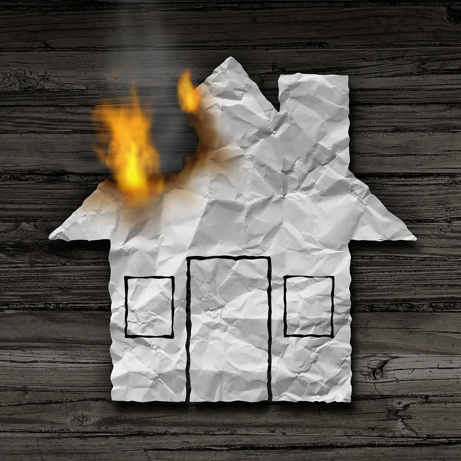 A crumpled white paper house cutout with drawn windows and door is burning on a dark wooden background.