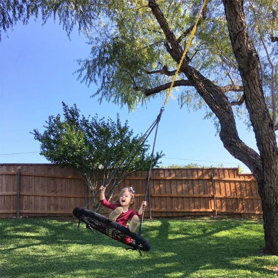 A joyful young girl laughs excitedly while swinging on a black saucer tree swing in a sunny backyard.