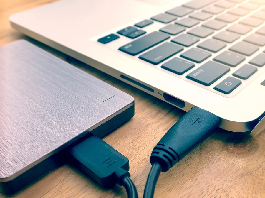 A brushed metal external hard drive connects to a silver laptop with a USB cable.