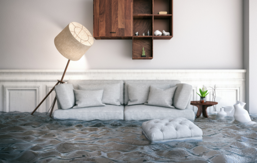 A flooded living room with a gray sofa, a tilted lamp, and water covering the floor, illustrating water damage.