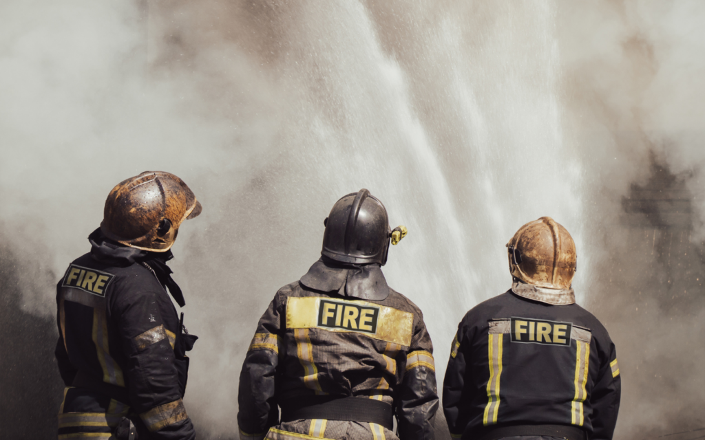 Three firefighters in protective gear stand with their backs to the viewer, facing a powerful torrent of