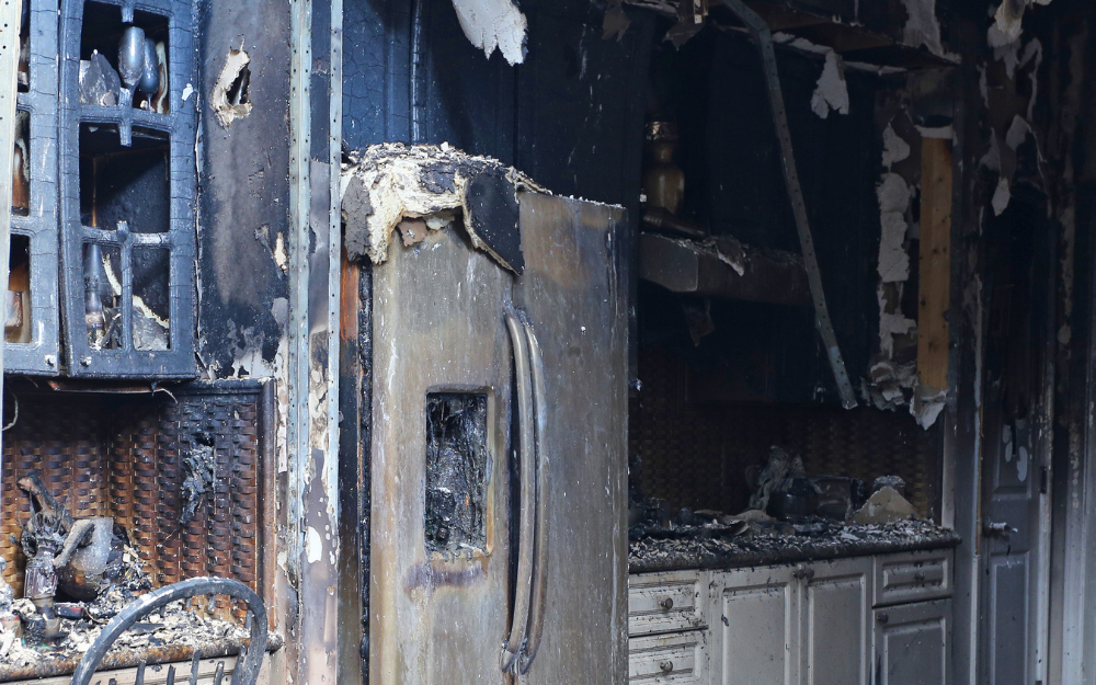 A severely fire-damaged kitchen features a burnt refrigerator, blackened cabinets, and debris.