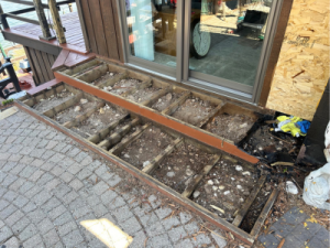 Partially deconstructed outdoor deck exposing damaged joists and substructure, in preparation for code-compliant reconstruction near a sliding glass door.