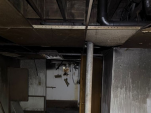 Basement mechanical area showing exposed ductwork, pipes, and a partially open door, with visible moisture staining and potential water damage on walls and ceiling—illustrating mitigation needs for dampness, mold risk, and structural protection.