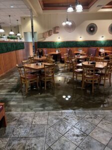 Restaurant dining area with tables and chairs on a partially flooded floor after a pipe burst, water spreading across the tile and carpeted sections, reflecting ceiling lights and creating standing water throughout the space.