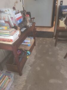 Cluttered room with a wooden table, books, and household items, showing carpet discoloration and damp spots caused by minor leaks near the doorway.