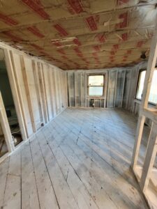 An interior view of a home restoration project featuring reconstruction and repairs due to fire damage. The room is gutted to the framework, highlighting structural wall studs, a wooden subfloor, a window, and freshly installed ceiling insulation.