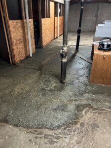 A wide shot of a basement floor heavily flooded with a thick, dark layer of wet sludge and dirty water. The murky residue spreads out across the concrete floor, showing swirling flow patterns. In the foreground, a vertical black drainpipe stands in the middle of the mess. To the left are wooden storage cubicles, to the right is the side of a wooden desk, and laundry machines are visible in the background.
