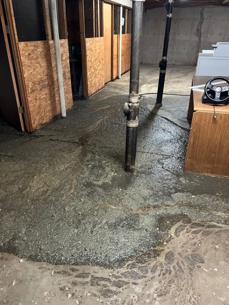 A wide shot of a basement floor heavily flooded with a thick, dark layer of wet sludge and dirty water. The murky residue spreads out across the concrete floor, showing swirling flow patterns. In the foreground, a vertical black drainpipe stands in the middle of the mess. To the left are wooden storage cubicles, to the right is the side of a wooden desk, and laundry machines are visible in the background.