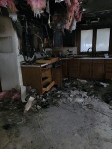 A devastating kitchen interior shows severe damage after a house fire, with charred walls and cabinets, debris covering the floor, and burnt insulation hanging from the ceiling.