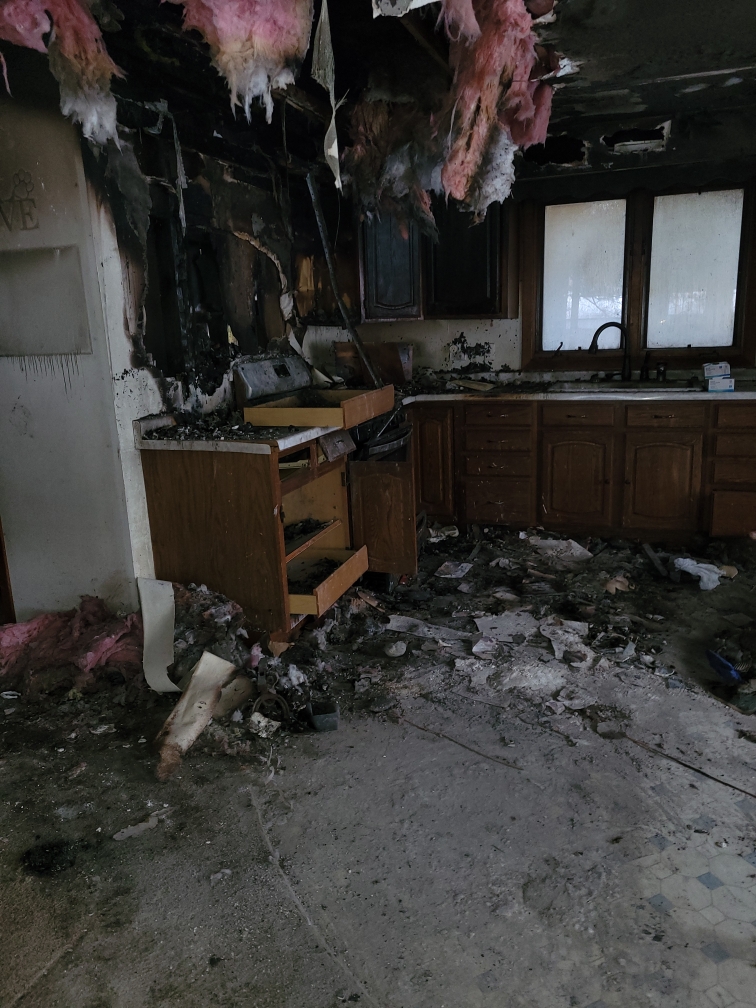 A devastating kitchen interior shows severe damage after a house fire, with charred walls and cabinets, debris covering the floor, and burnt insulation hanging from the ceiling.
