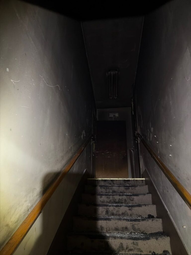 Dark narrow staircase showing severe smoke damage, heavy soot accumulation, and fire residue on the walls and steps leading up to a door.