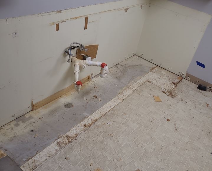 Exposed subfloor water damage and dark mold staining underneath a leaking sink pipe after cabinet removal.