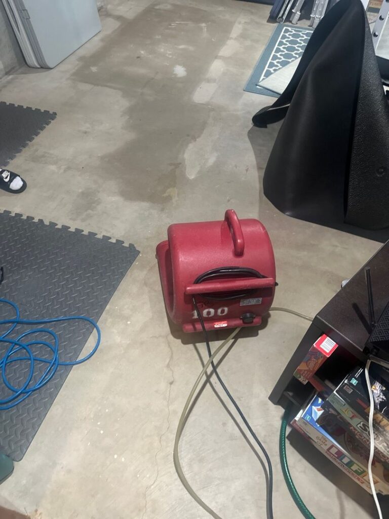 Red commercial air mover and hoses drying a concrete floor after an appliance leak in a basement utility area.