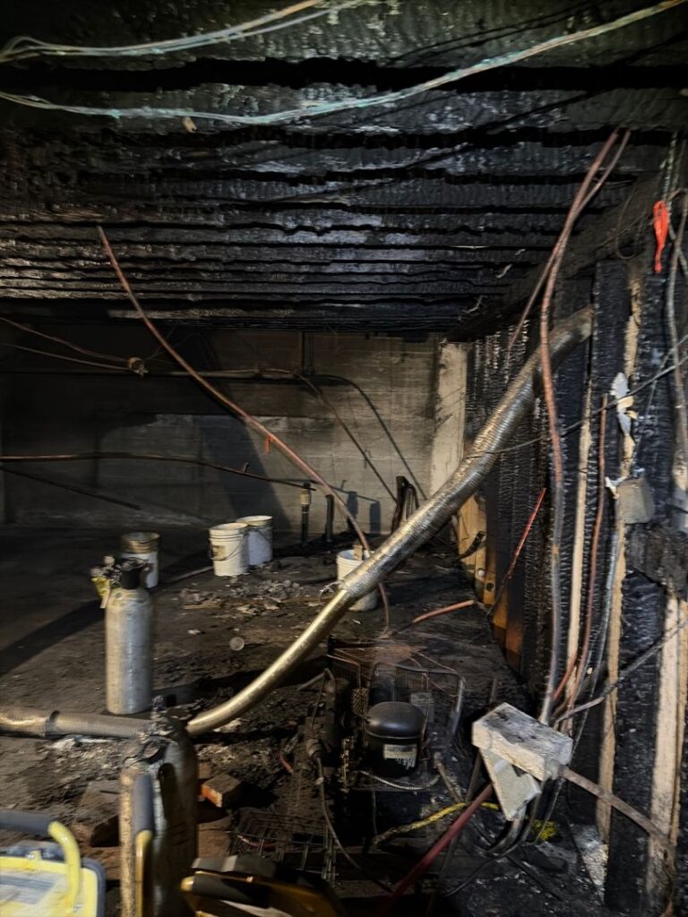 Severely fire-damaged basement room showing exposed electrical wiring, charred wooden beams, and burnt equipment.