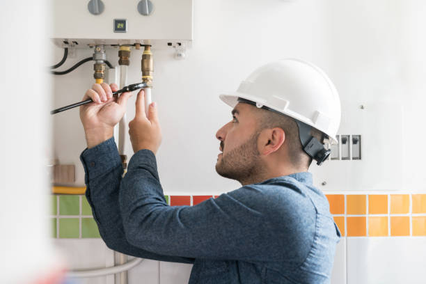 A plumber in a white hard hat uses a wrench to service a wall-mounted water heater.