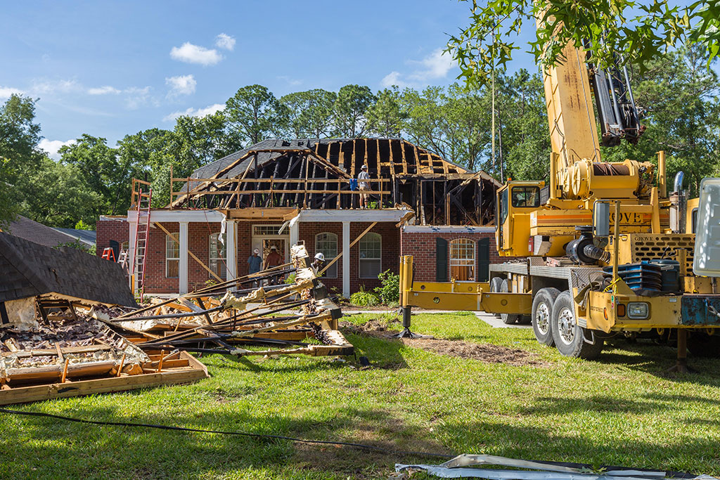 Construction site with a partially roofed house, a crane, and debris scattered on the lawn under a clear blue sky.