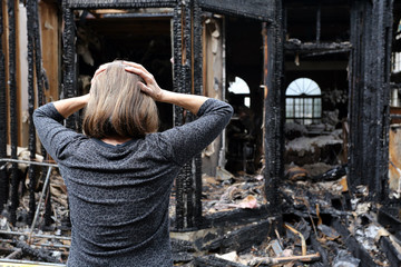 A woman stands with her hands on her head, looking at the charred remains of a house after a fire.