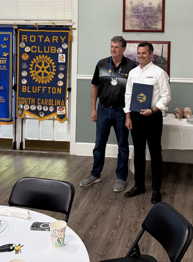 Smiling men stand before a Rotary Club Bluffton banner, one holding a presentation folder.
