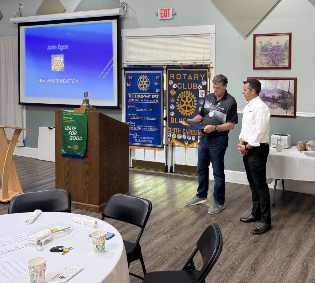 John Egan is inducted as a new Rotary Club member, standing with another man near club banners.