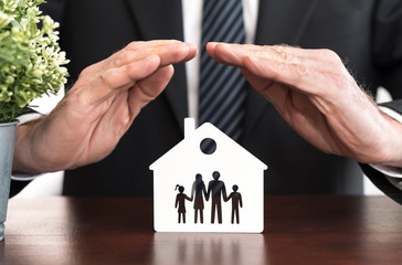 A professional's hands cup over a house and family cutout, symbolizing protection and