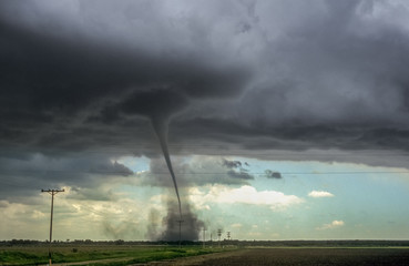 A powerful tornado descends from dark storm clouds over a flat, open landscape with utility poles
