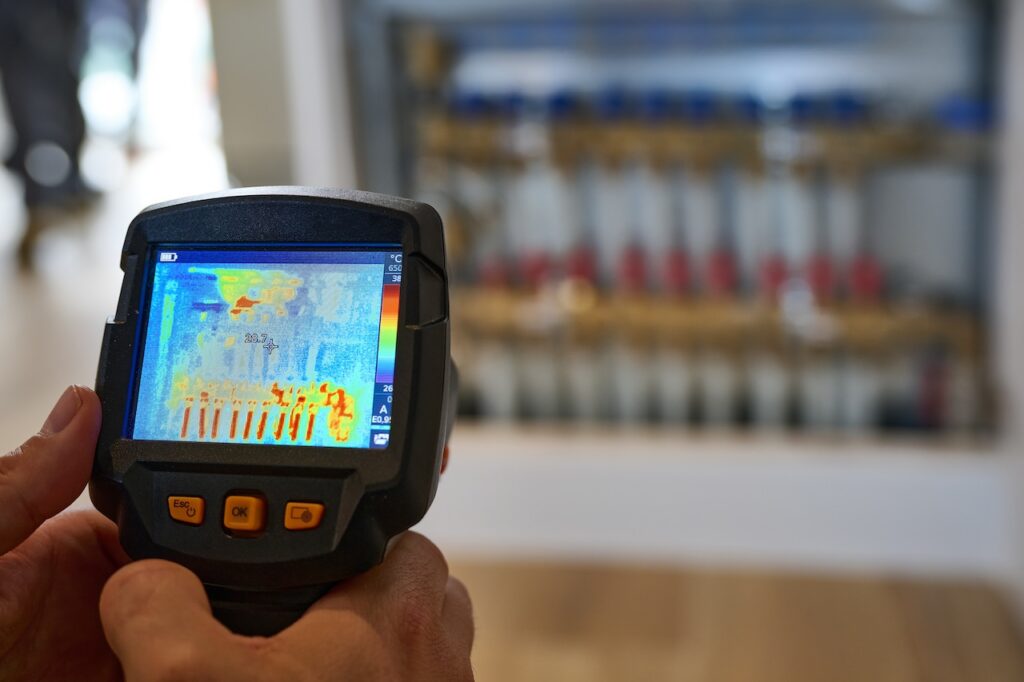 Puroclean of Huntington checking for an underground water leak with a thermal imaging tool. The image depicts a handheld thermal camera being used to measure the temperature of an underfloor heating manifold. The camera is held in both hands, and the operator is pointing it directly at the manifold