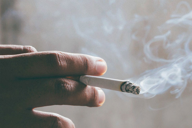 A close-up image of a person's hand holding a lit cigarette, with wisps of smoke rising against a dark background. The burning tip of the cigarette glows slightly, leaving behind a trail of ash. The smoke creates a hazy effect, emphasizing the lingering smell that can cling to skin, clothing, and indoor spaces.