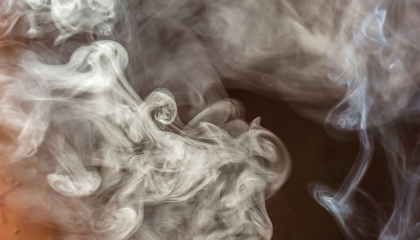 A close-up image of swirling white smoke against a dark background, creating intricate, fluid patterns as it rises and disperses. The tendrils of smoke form delicate curls and wisps, giving a sense of movement and depth. The image evokes the lingering, often pungent smell of smoke, which can cling to surfaces and fabrics long after the source has disappeared.