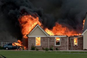 A single-story brick home engulfed in flames with heavy black smoke rising into the sky; the roof and interior are burning intensely while a nearby car is also on fire, illustrating severe residential fire damage.