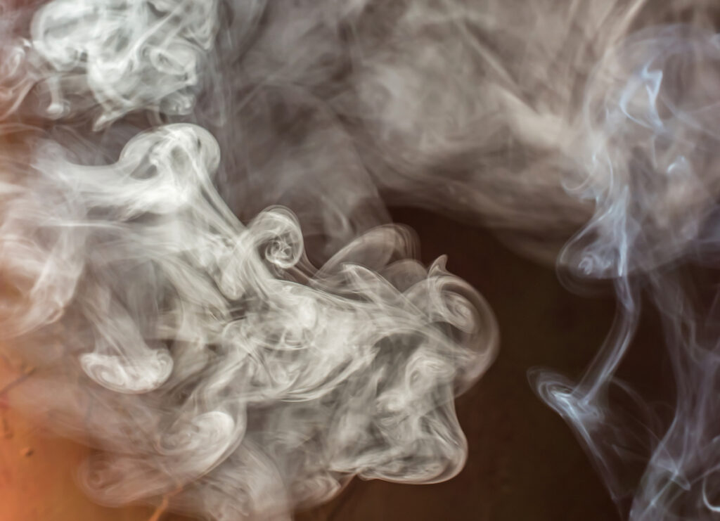 A close-up image of swirling white smoke against a dark background, creating intricate, fluid patterns as it rises and disperses. The tendrils of smoke form delicate curls and wisps, giving a sense of movement and depth. The image evokes the lingering, often pungent smell of smoke, which can cling to surfaces and fabrics long after the source has disappeared.