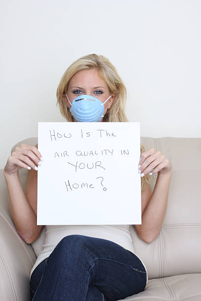 Woman wearing a blue face mask holds a sign asking 'How Is The Air Quality In Your Home