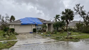 Hurricane-damaged house with a blue tarp covering its roof and debris scattered across the yard.