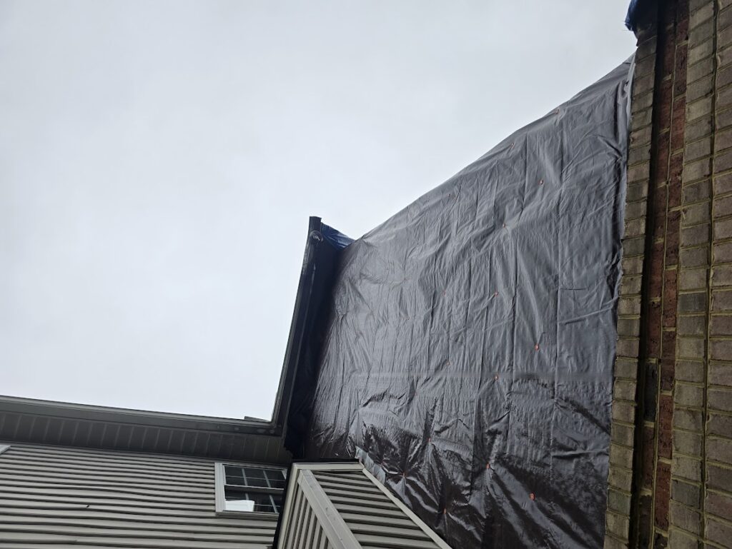 A house roof is covered with a black tarp next to a brick wall under an overcast sky.